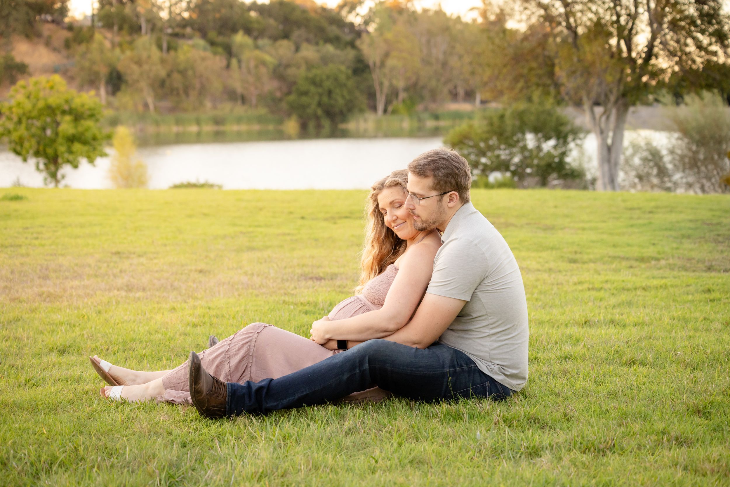 A couple sits on the grass with lake in the background for their maternity photo session