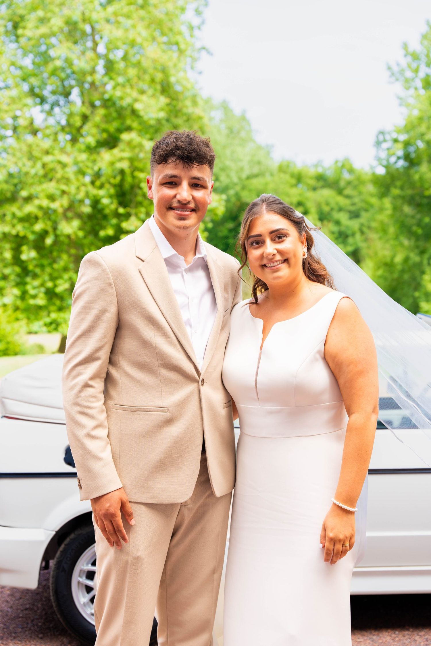 A couple poses together outdoors in formal beige and white attire.