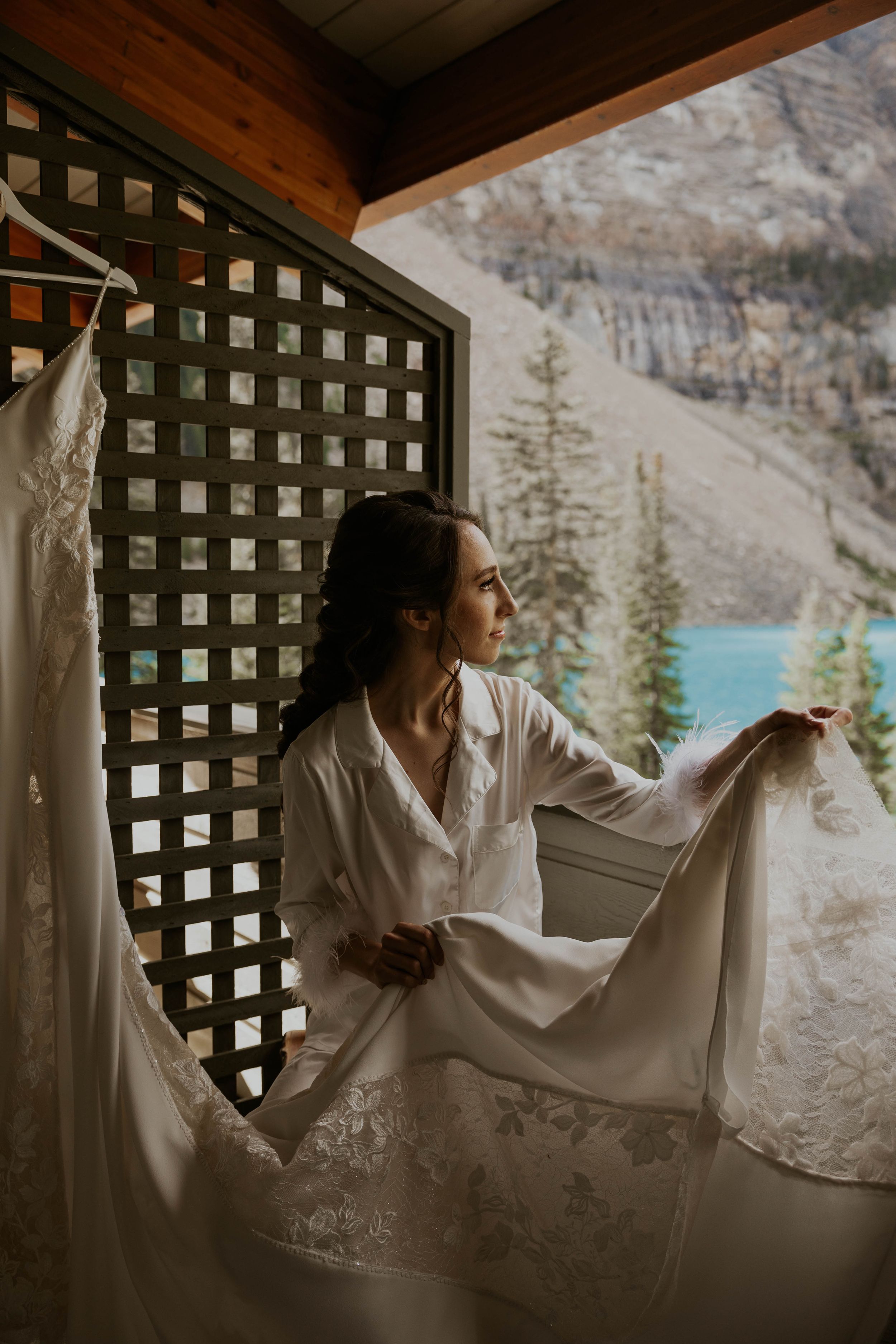 White wedding dress hanging in rustic cabin overlooking turquoise mountain lake and rocky cliffs.