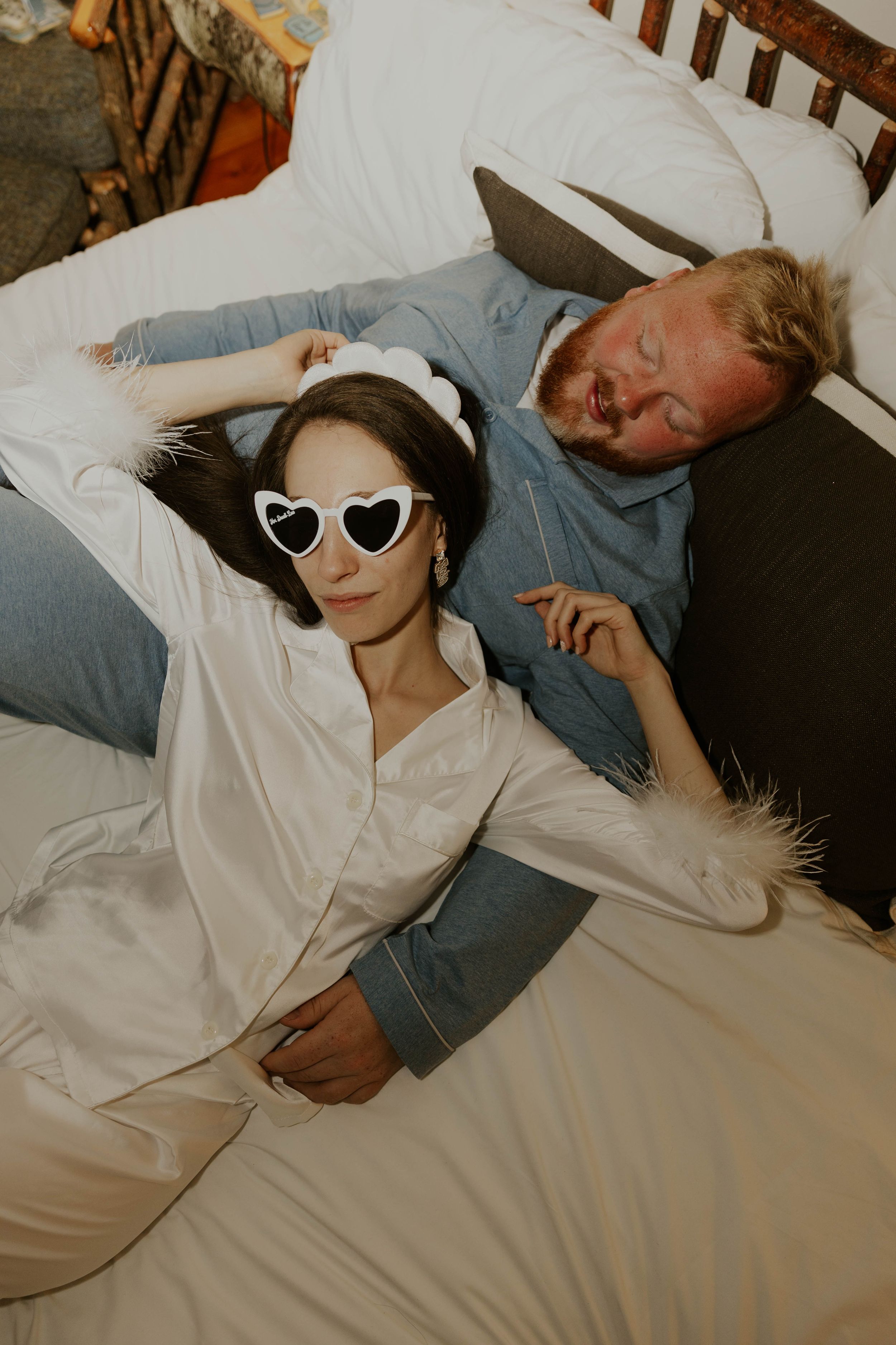 Two people relax on white bedding, one wearing heart-shaped sunglasses.