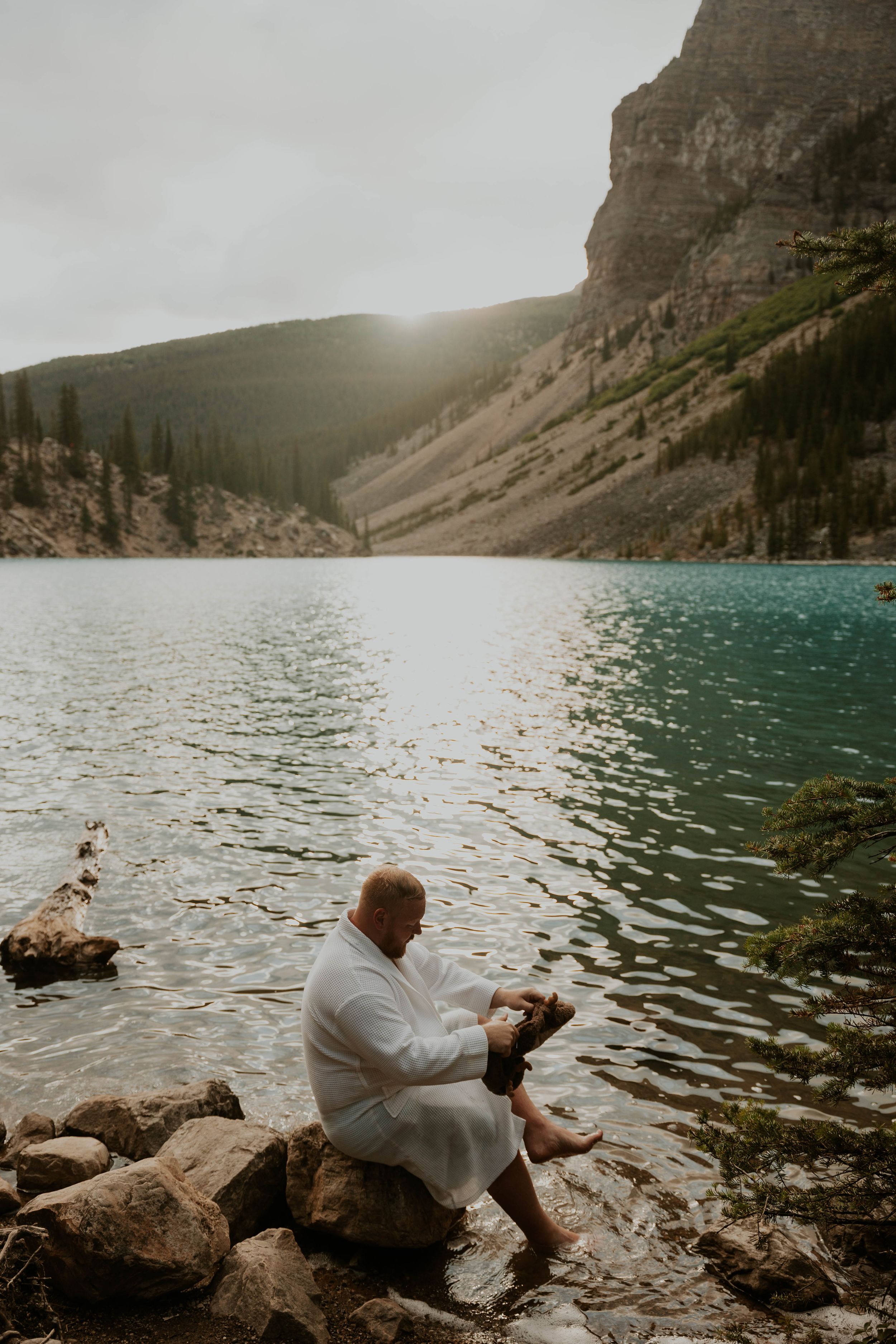 A person in white sits on rocky shoreline beside a mountain lake at sunset.