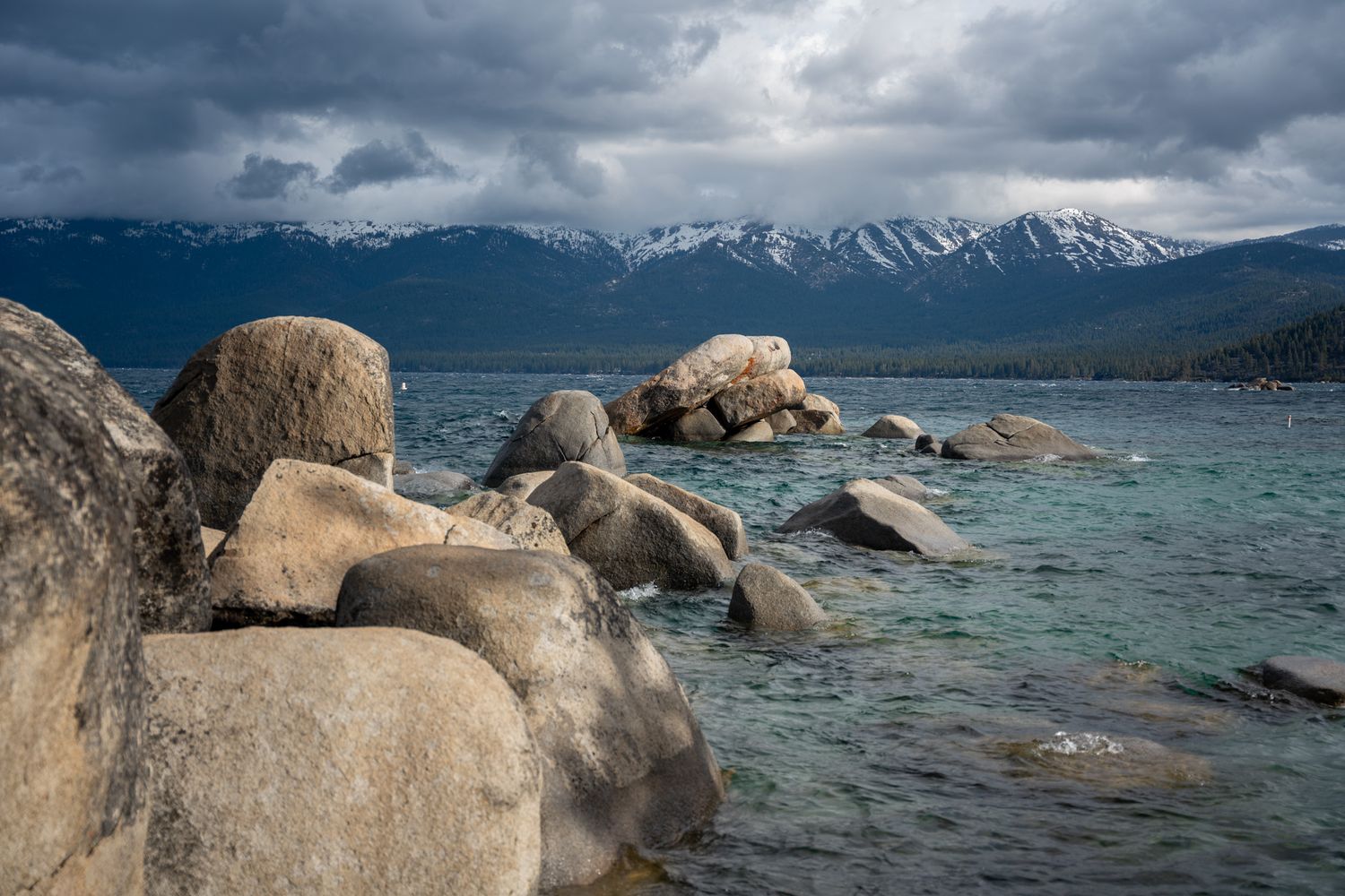 Large boulders dot the shoreline of a mountain lake with snow-capped peaks in the background.