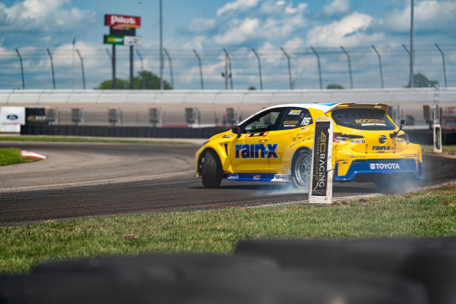 A yellow rally car drifting on a racetrack with tire smoke trailing behind.
