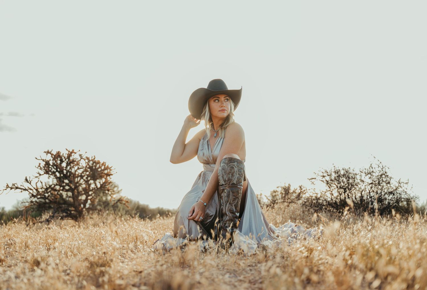 A person in a cowboy hat and flowy dress sits in a golden sun-drenched field during sunset.