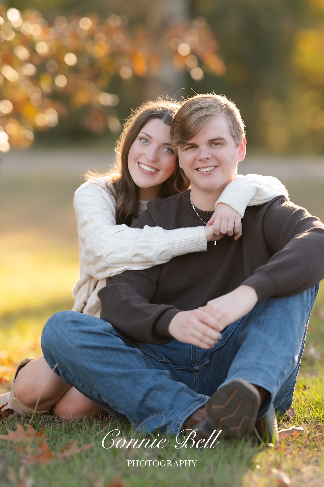 Couple sitting together on grass during golden hour with autumn bokeh lights in background.
