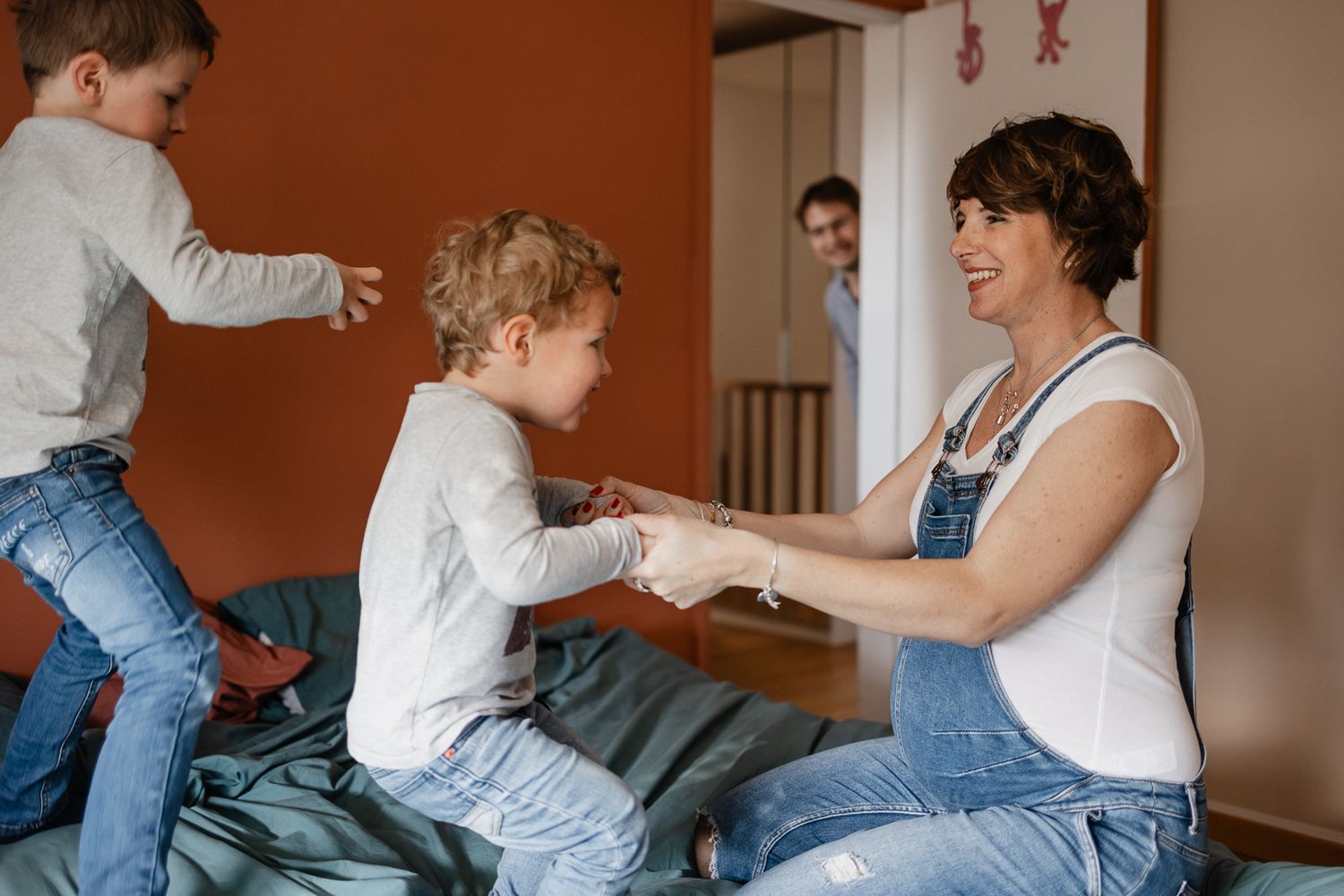 photographe lille : maman attendant son 3e enfant et jouant sur le lit avec ses deux aines, le papa observe la scene amuse en arriere plan