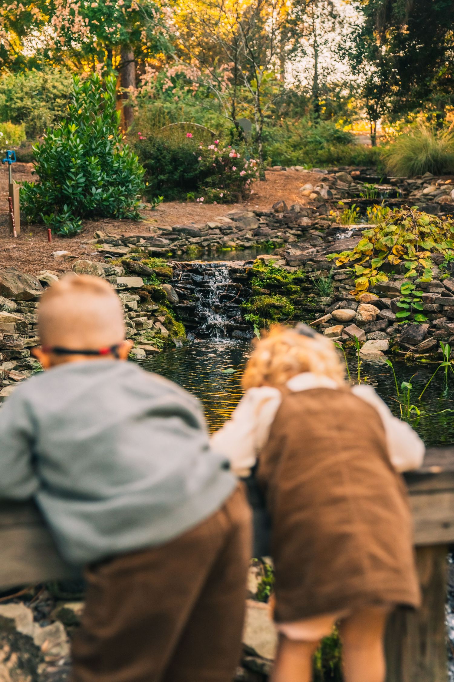 Two people sit together enjoying a peaceful view of a stone waterfall feature in a garden at sunset.