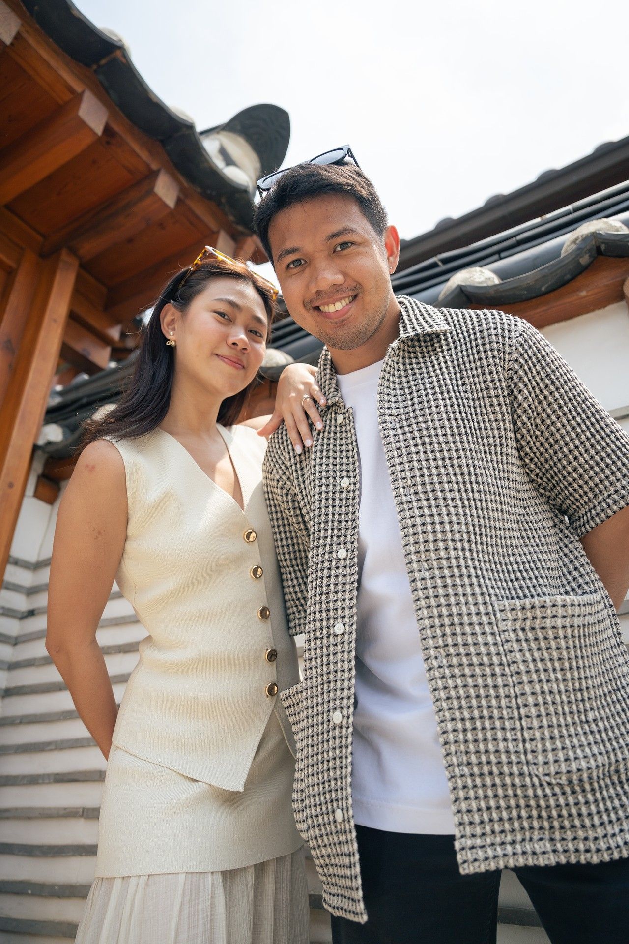 A couple poses together outdoors wearing coordinating outfits of a cream dress and checkered jacket.
