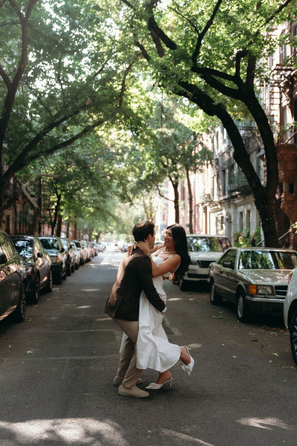 A romantic couple dances together on a tree-lined city street with parked cars and dappled sunlight.