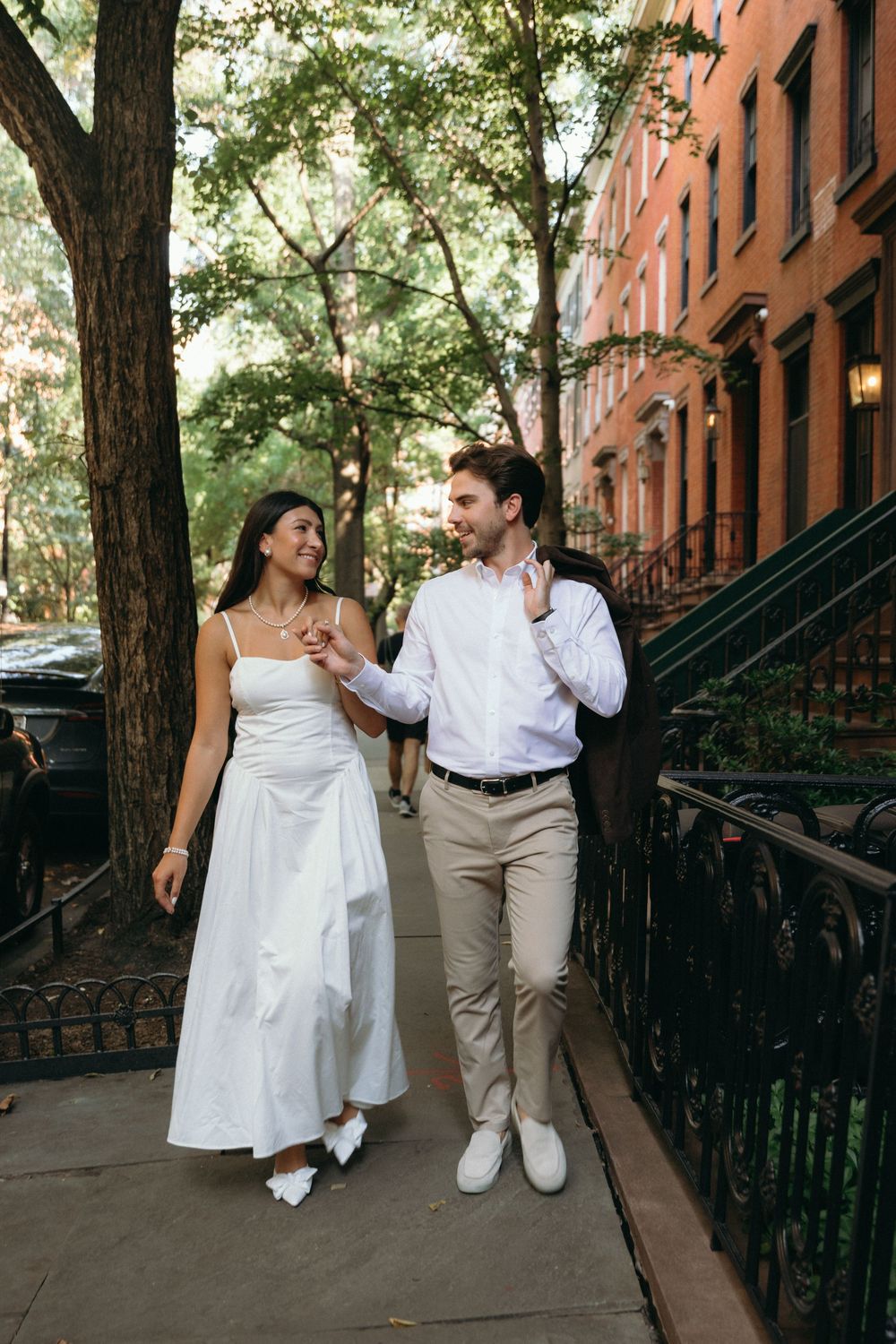 A couple walks together on a brownstone-lined street in summer, with the person in white wearing a flowing dress.