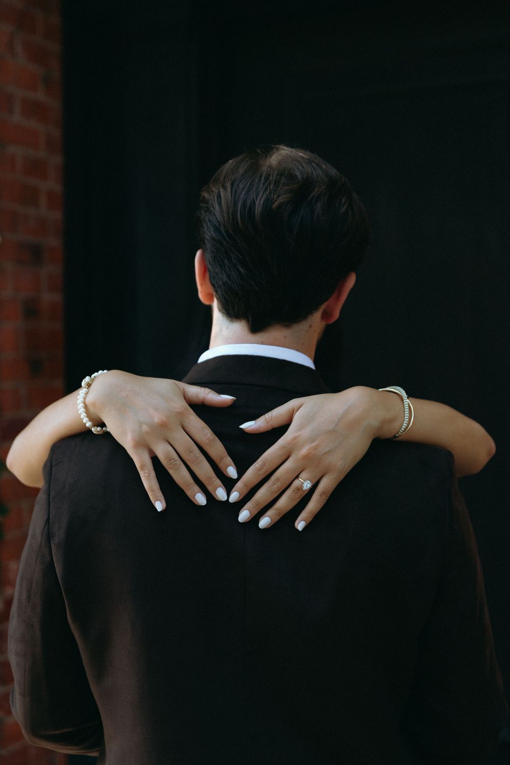 A close-up view of hands embracing someone wearing a black suit jacket against a dark background with brick wall.
