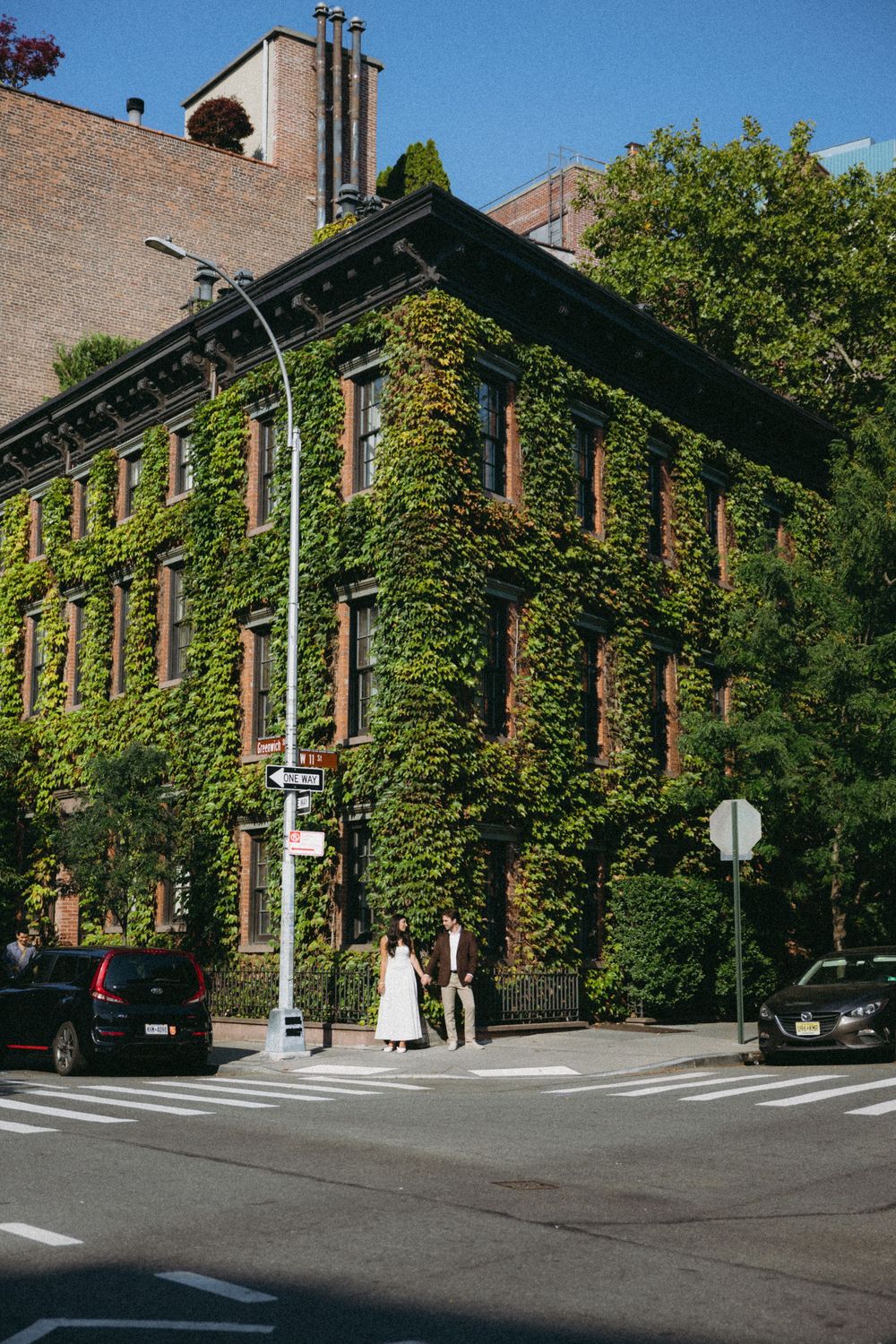 Brick building with ivy-covered walls and trees along a city street corner on a sunny day.