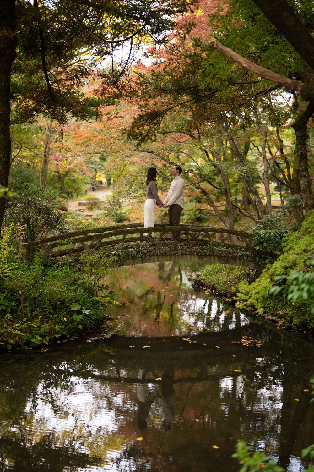 Couple stands on an arched stone bridge reflected in calm water amid lush autumn trees in a serene Japanese garden.