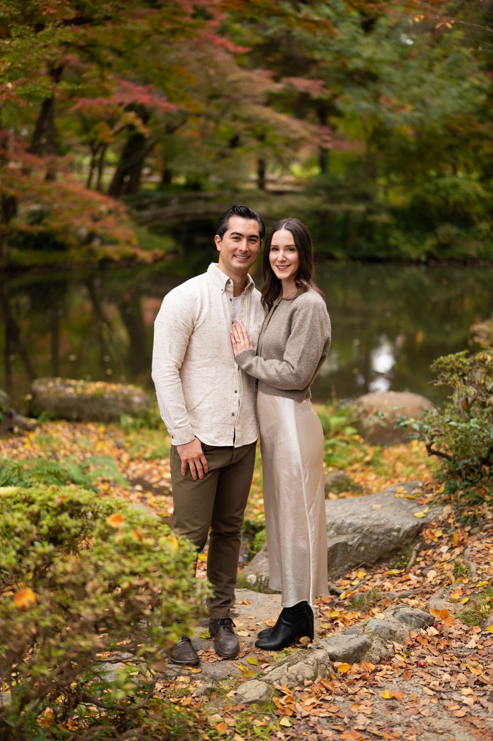 Smiling couple poses together on a stone path beside a pond with vivid autumn trees in a Japanese garden.