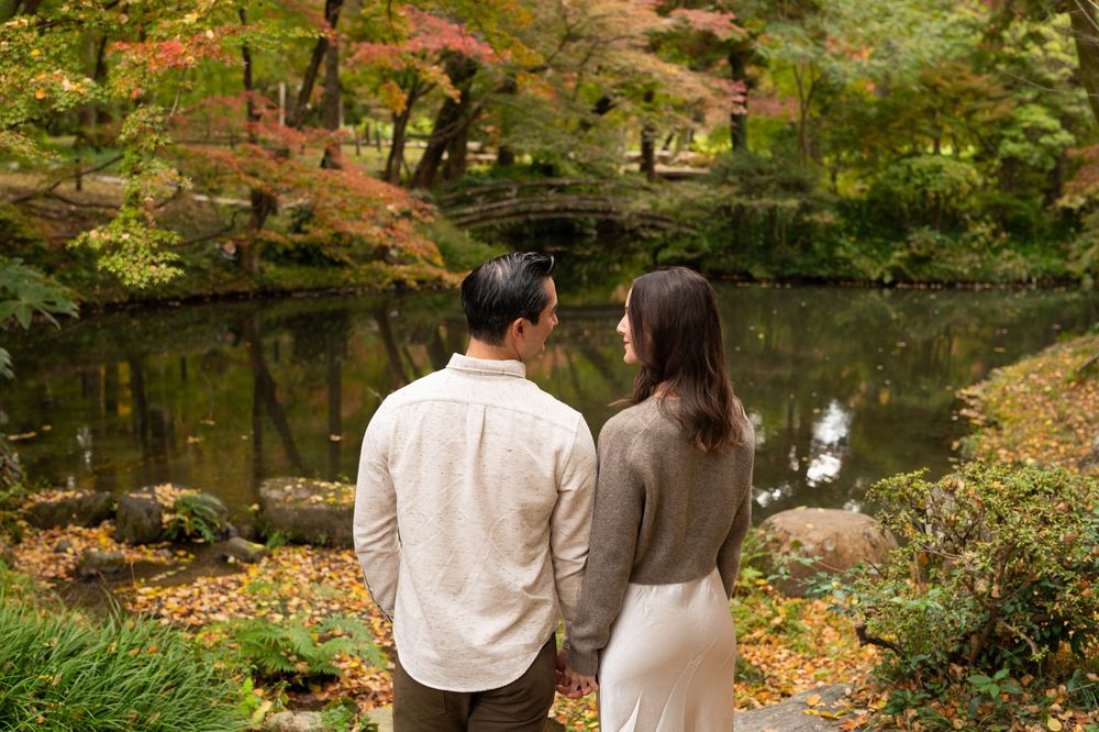 Couple standing hand in hand by a tranquil Japanese garden pond surrounded by colorful autumn foliage.