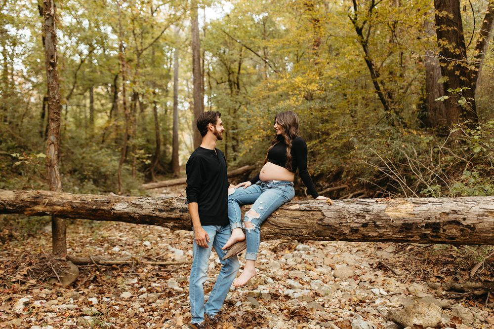 A person in blue jeans sits on a fallen log in an autumn forest with golden leaves scattered on the ground.