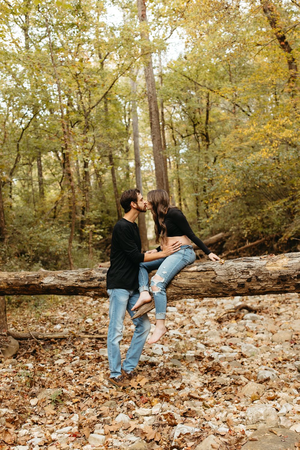 A couple playfully dances in a wooded area covered in fallen autumn leaves.