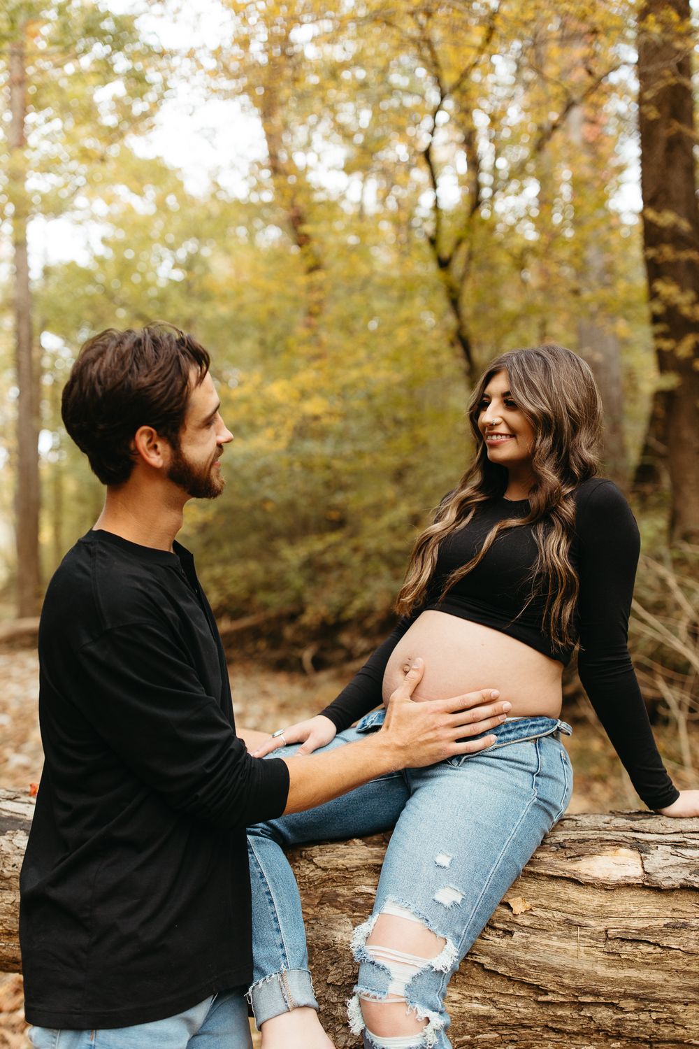 A couple playfully interacts while sitting on a fallen tree trunk in an autumn forest setting.