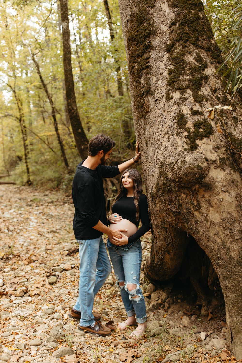 Two people share an intimate moment against a large tree trunk surrounded by fallen autumn leaves.