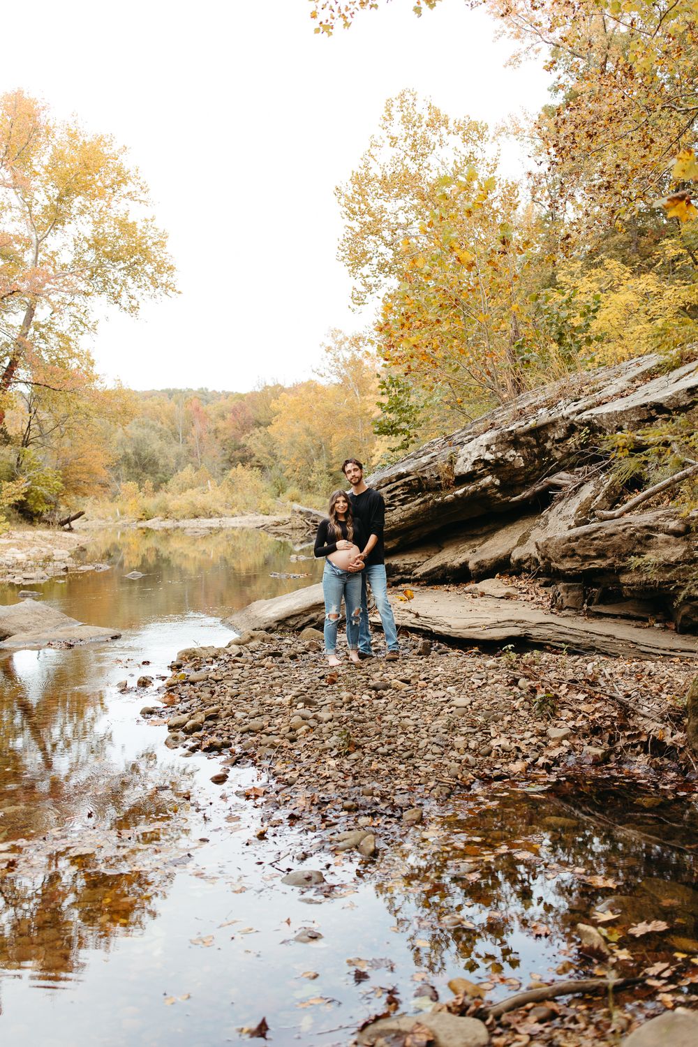 A couple stands by a rocky riverbank surrounded by autumn trees and fallen leaves reflecting in calm waters.