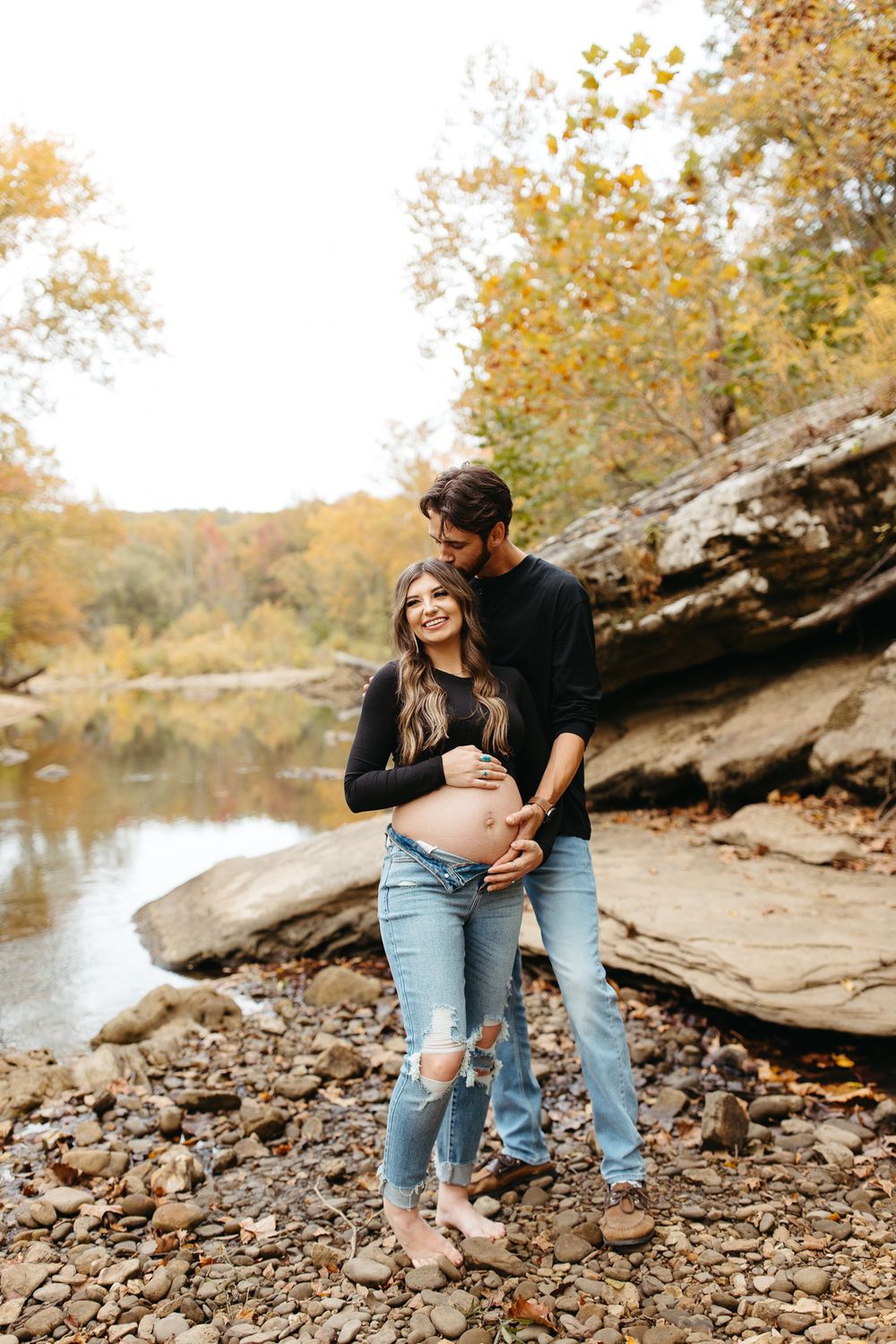 A couple embraces by a rocky riverbank surrounded by autumn trees with golden foliage.