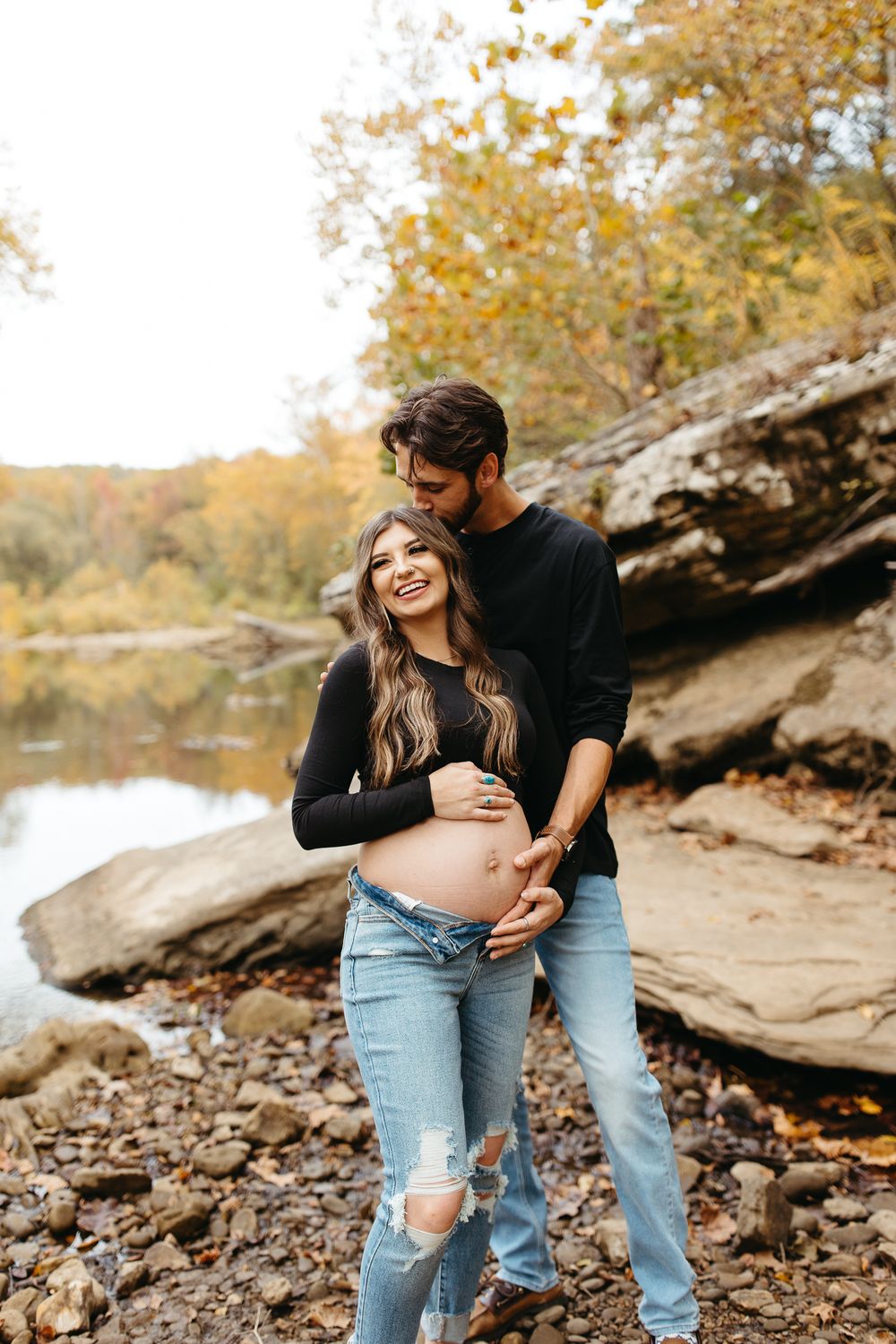 Couple embraces while sitting on rocky shoreline during autumn with water and fallen leaves visible.