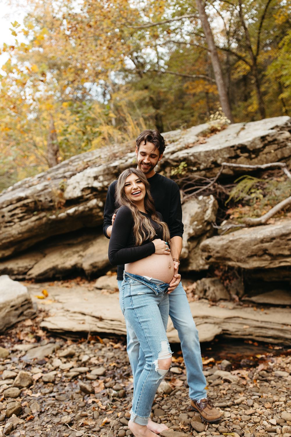 Romantic couple shares intimate moments on rocky ground with fall trees and golden leaves in background.