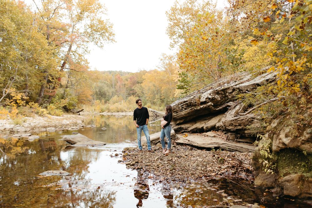 A couple walks along a scenic creek with rocky outcrops and autumn trees.