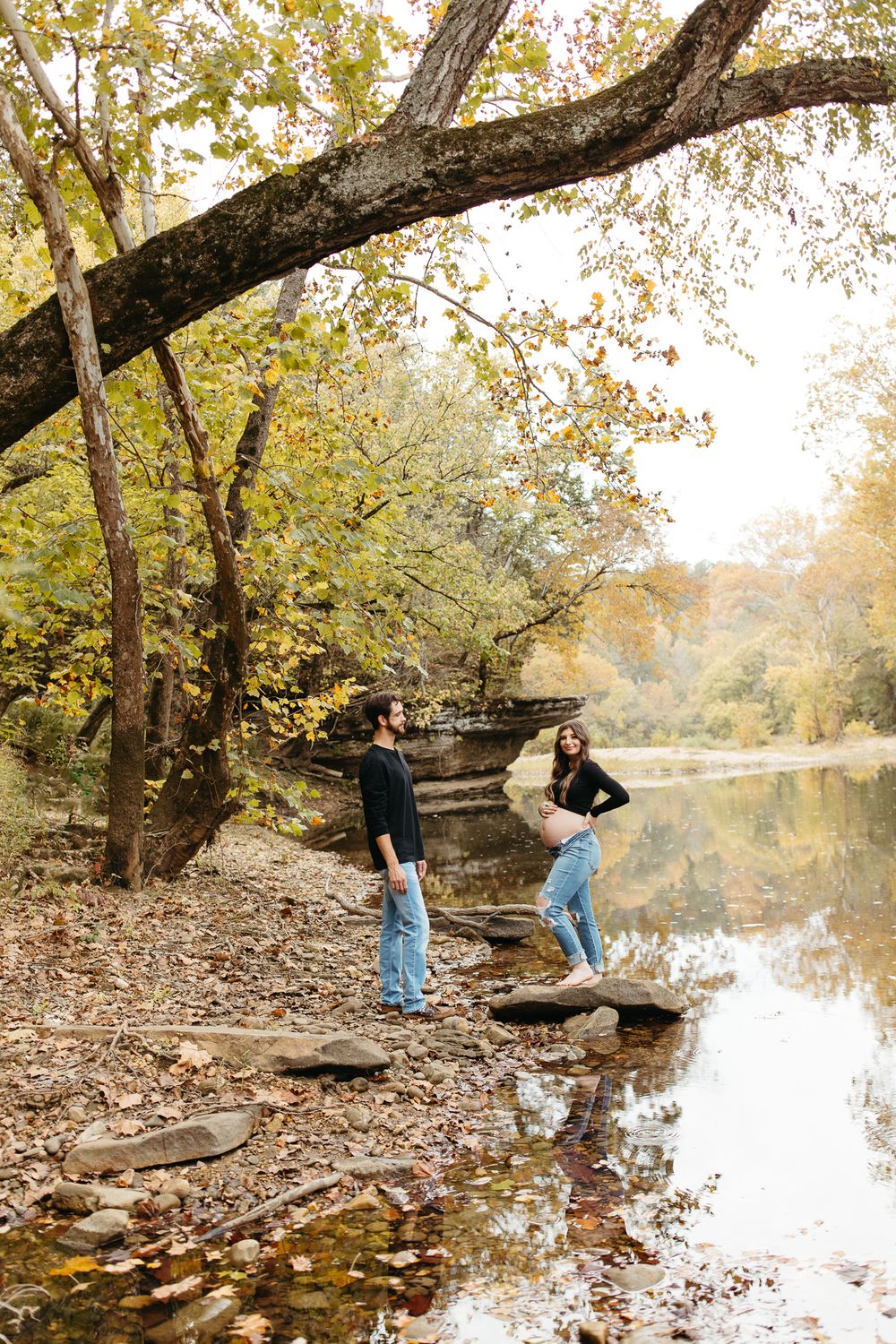 Two people stand on rocky shoreline by tranquil pond surrounded by autumn trees in golden afternoon light.