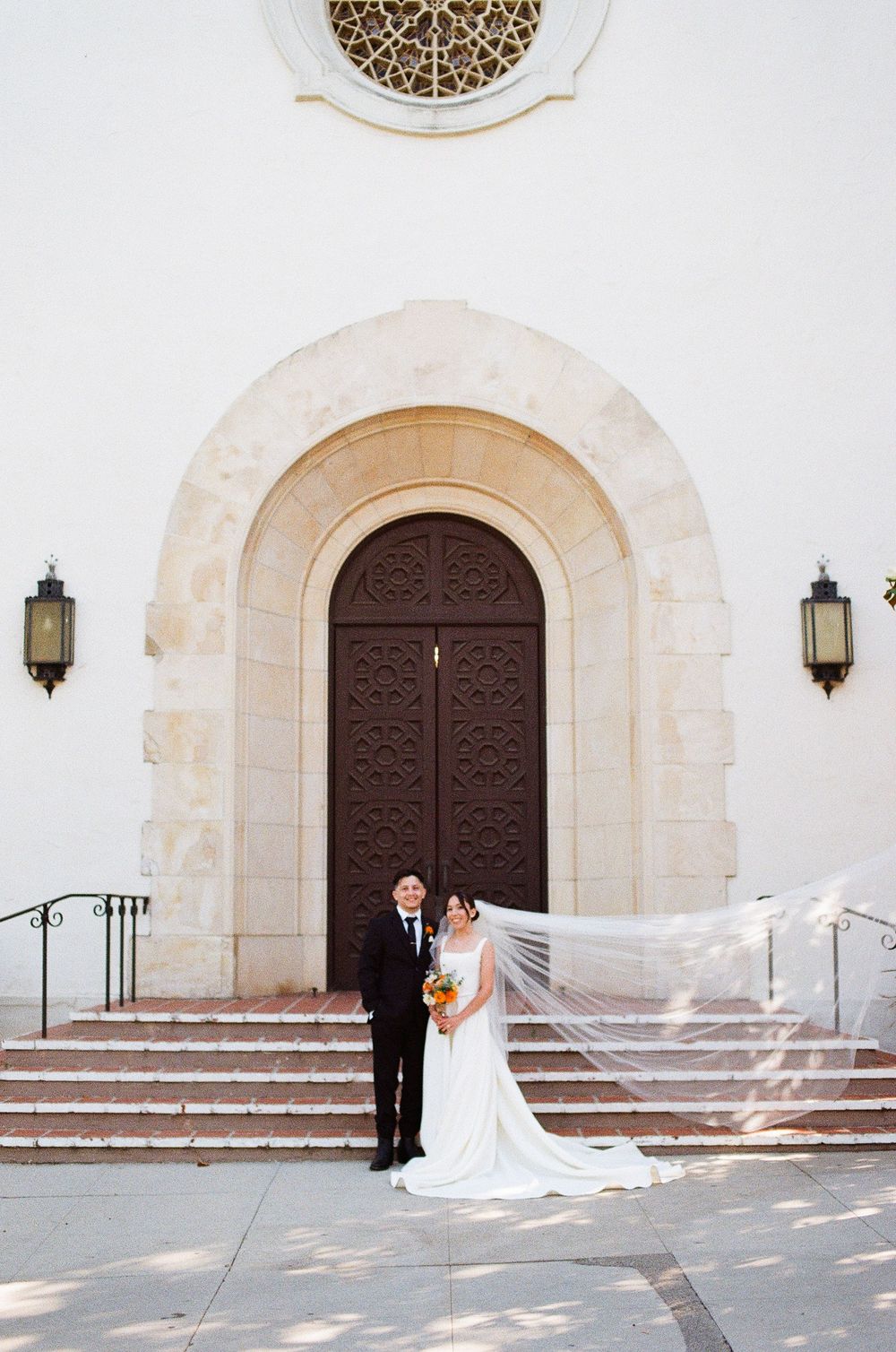 Film photography in Santa Barbara of a bride with flowing veil and groom in black tuxedo in front of chapel
