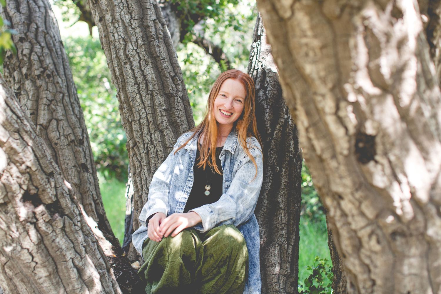 Person in denim jacket poses against tree trunk in natural outdoor setting.