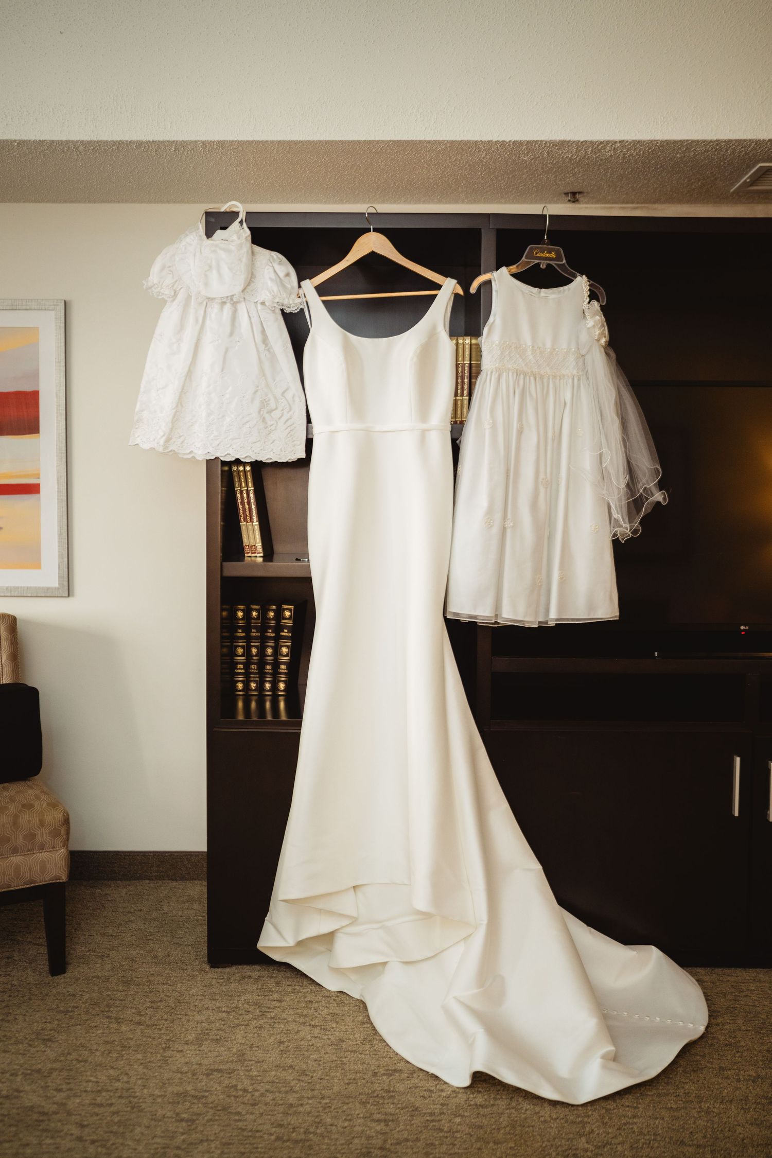 White wedding dresses and flower girl dresses hanging on wooden hangers in a hotel room before the ceremony.