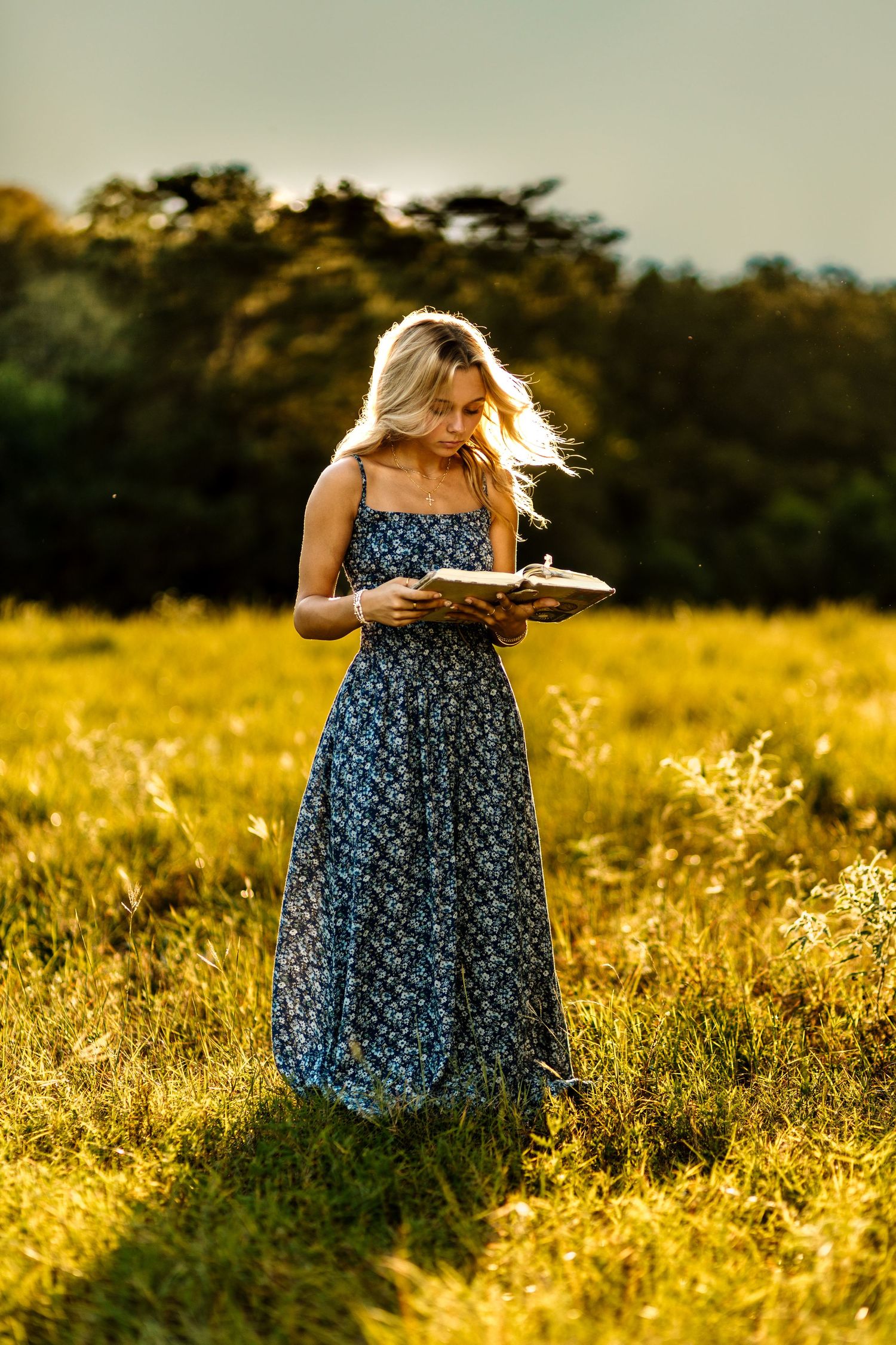 Person in a blue floral dress stands in a golden meadow at sunset with tall grass and wildflowers swaying in the breeze.