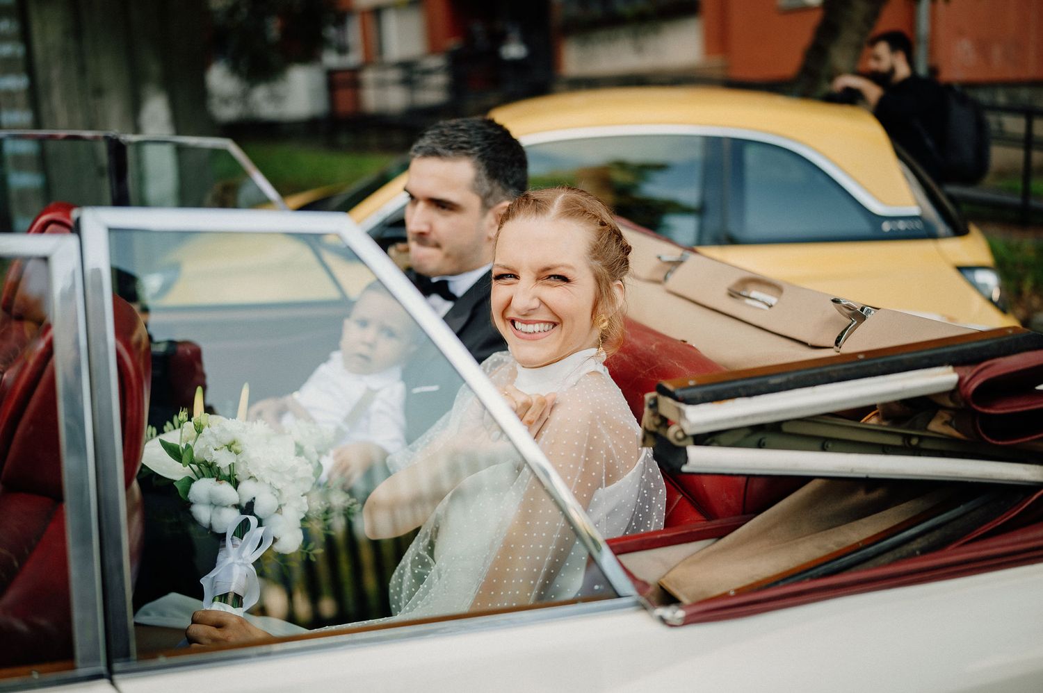 A happy couple rides in a vintage red convertible car on their wedding day with white floral bouquet visible.