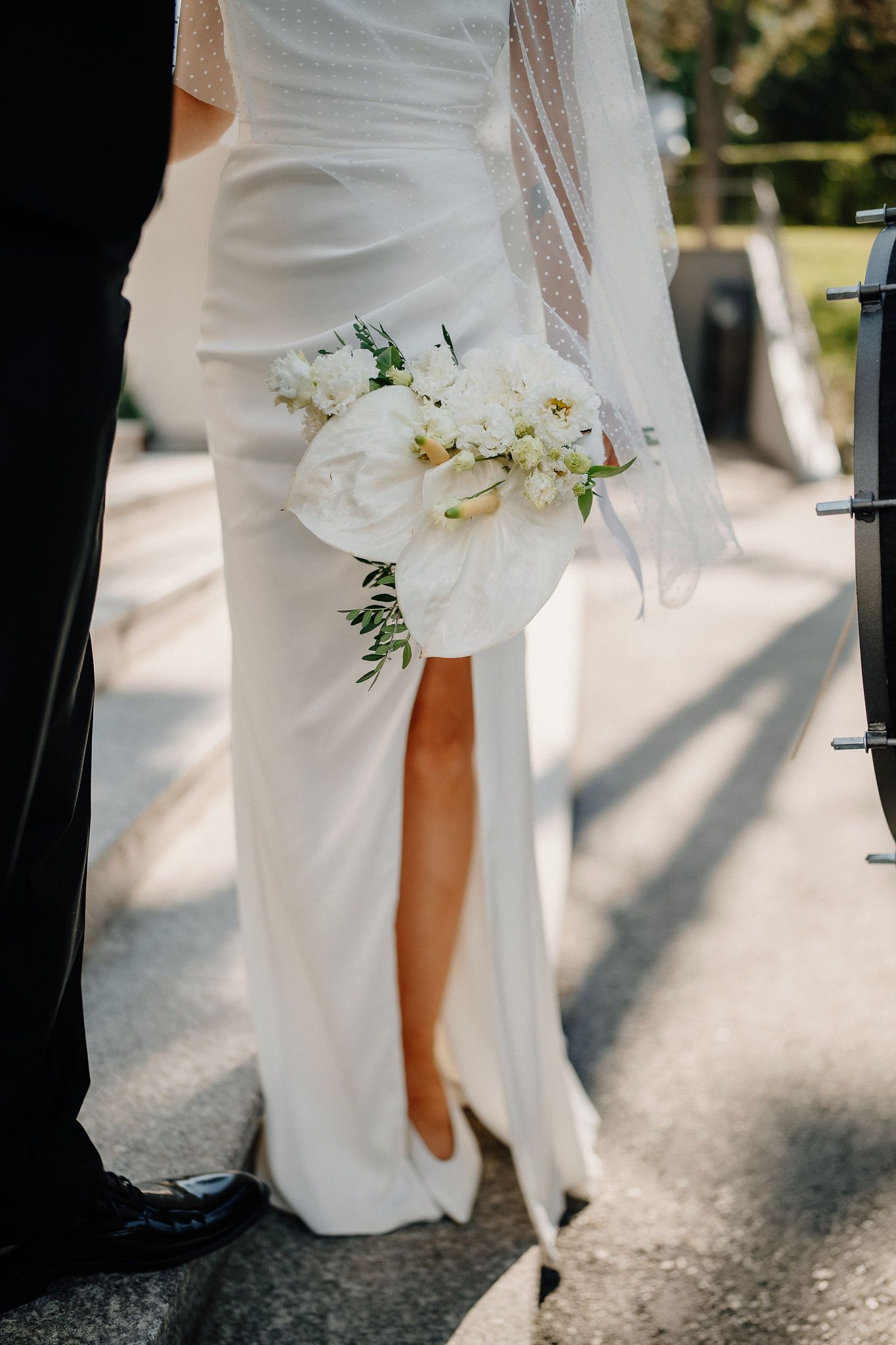 Elegant white wedding dress detail with flowing skirt and delicate white orchid bouquet against sunlit pavement.