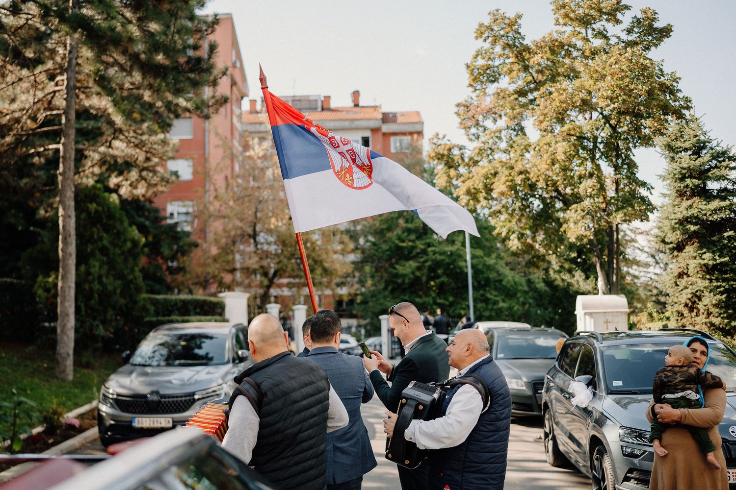 Serbian flag waving during an outdoor celebration with parked cars in the background.