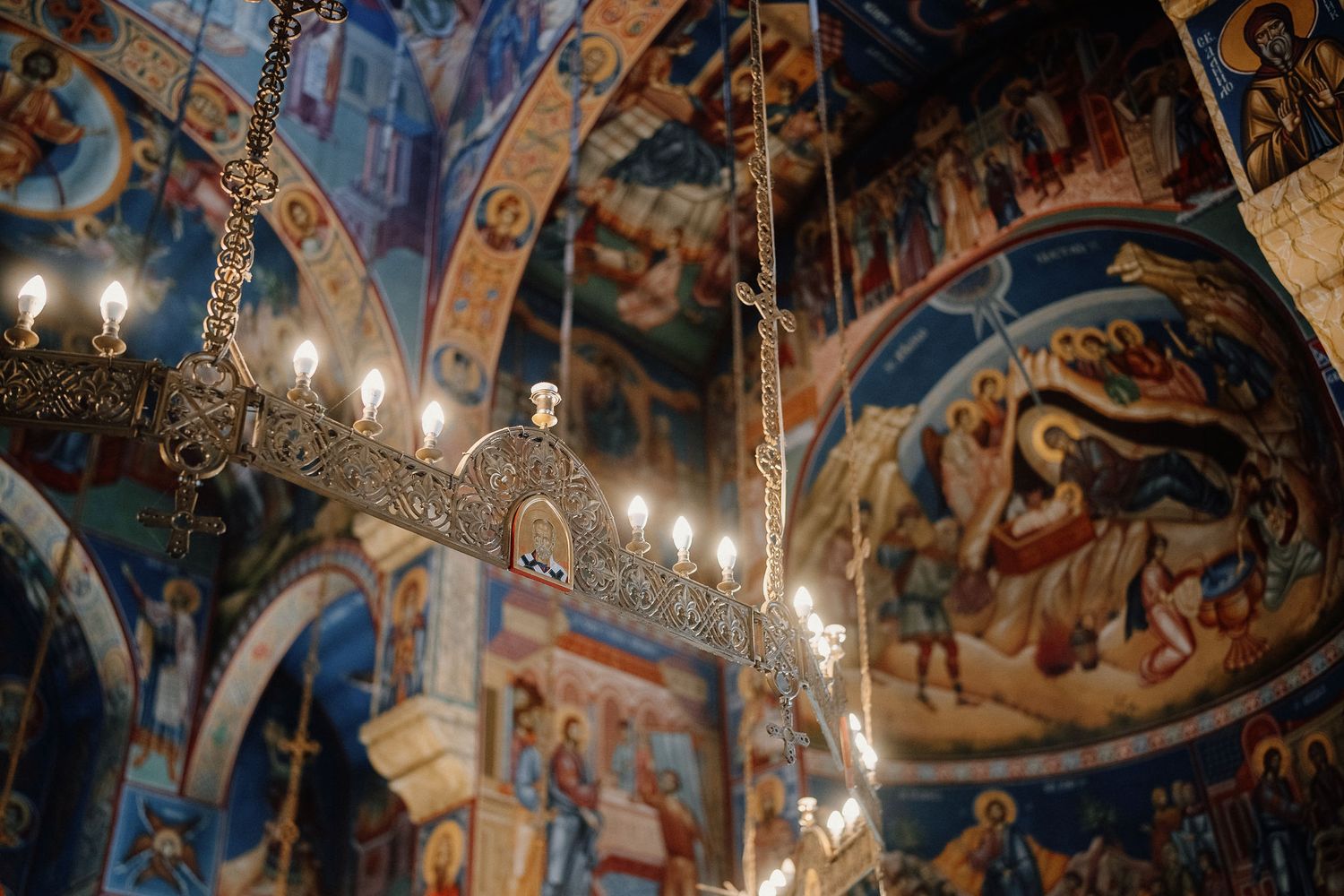 Ornate Byzantine church interior with religious frescoes covering walls and arches, illuminated by hanging chandeliers.