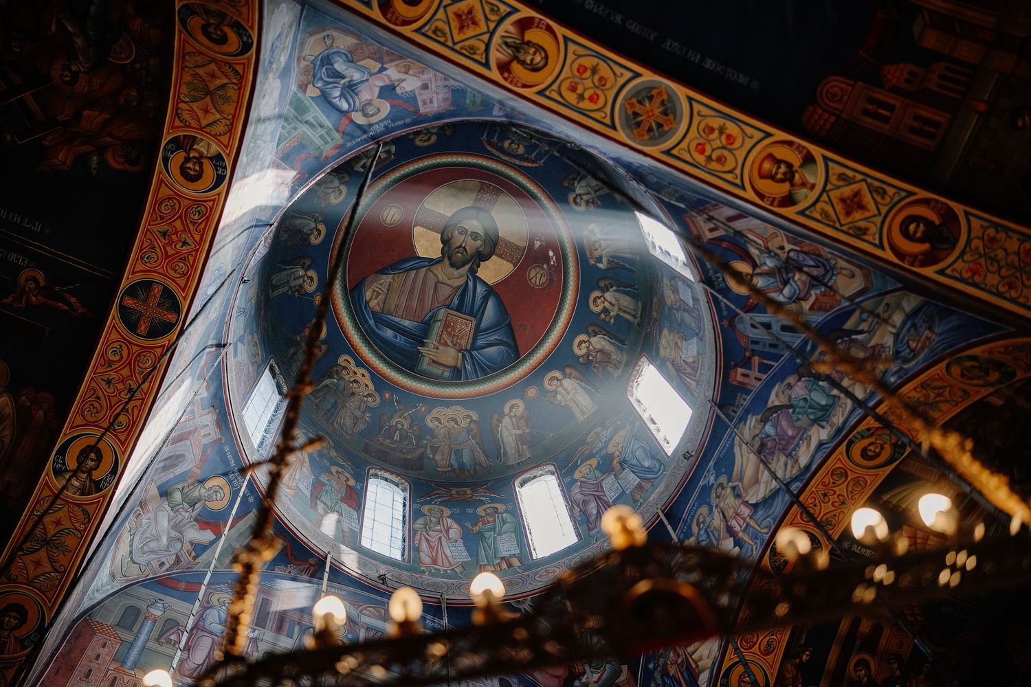Ornate Byzantine church interior with golden mosaics and religious frescoes illuminated by hanging pendant lights.