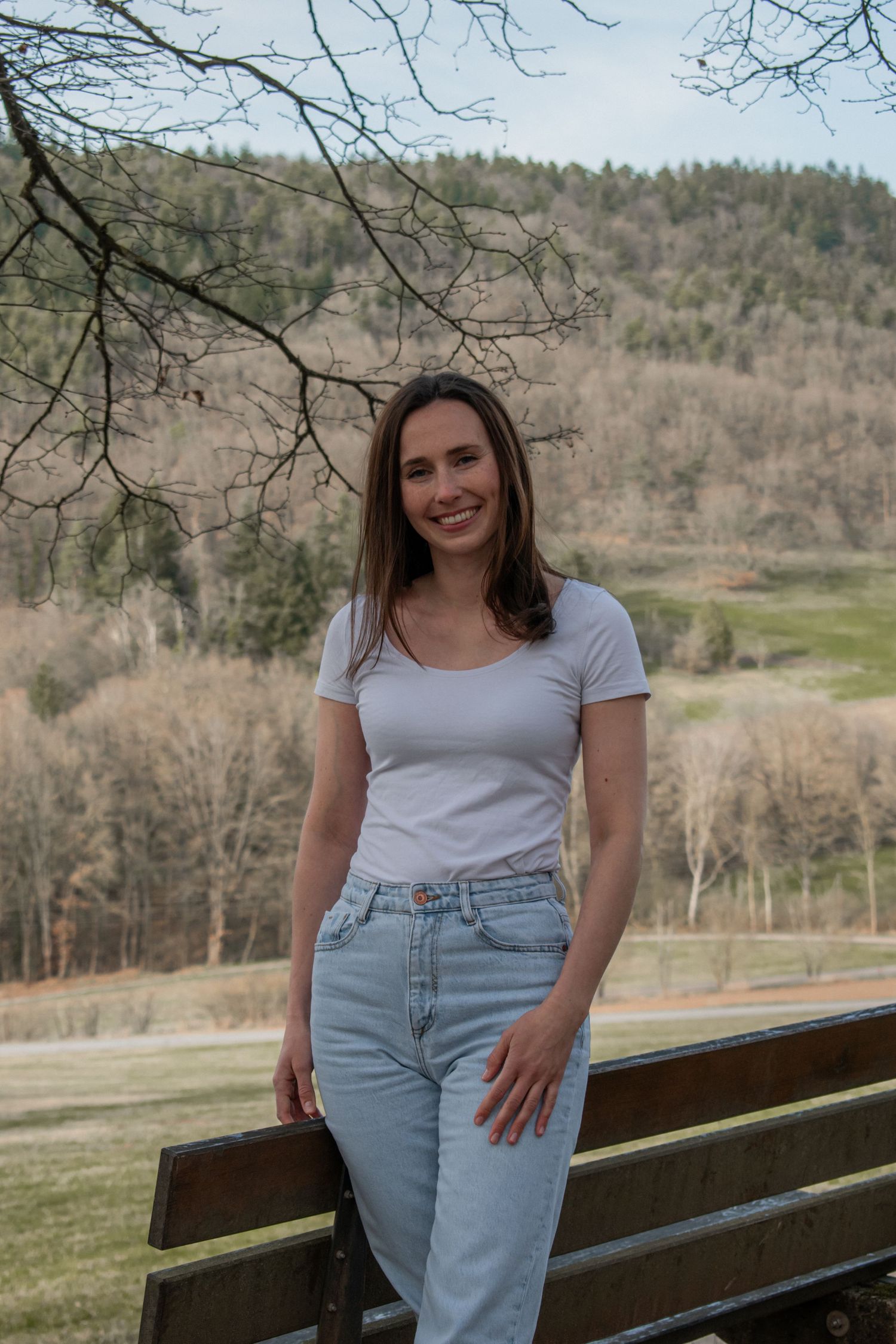 Casual portrait in nature showing someone in a white t-shirt and light jeans leaning against wooden railing with hills behind.