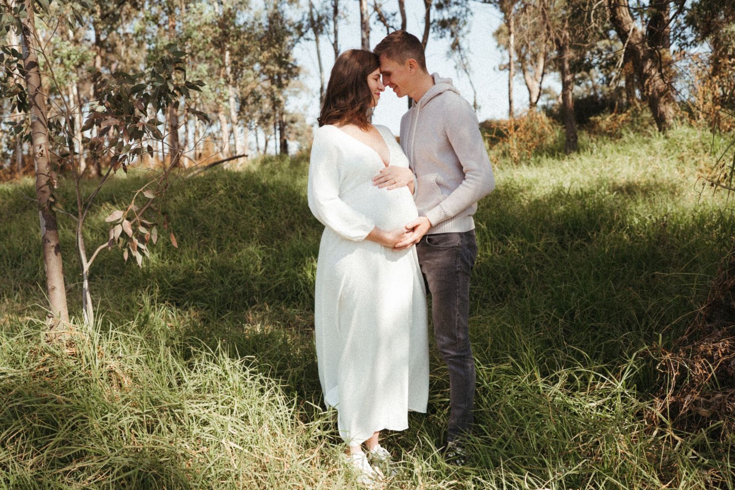 Romantic couple embracing in sunlit wooded area wearing white and grey casual attire.