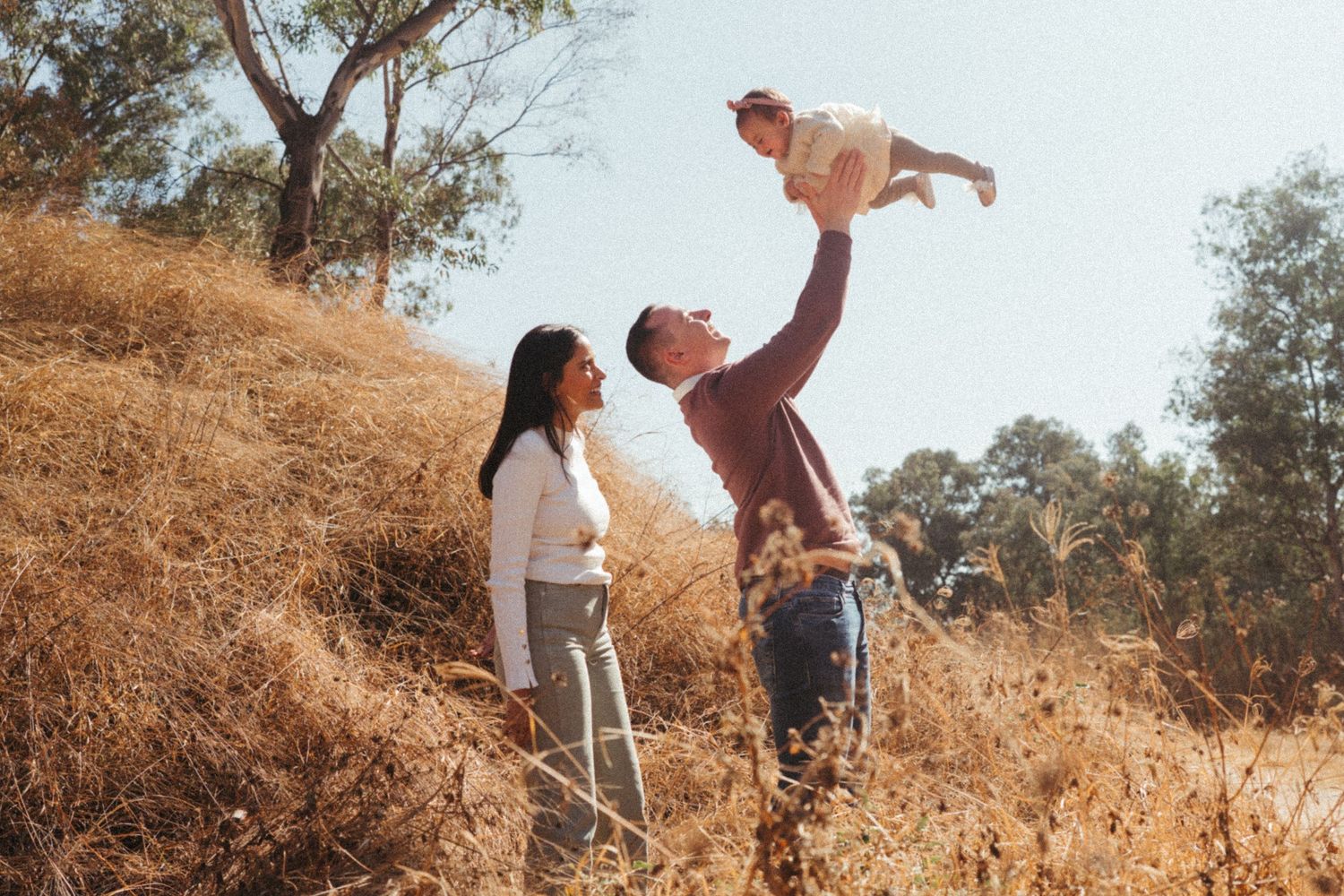 Playful family interaction on a sunlit hillside with dried vegetation.