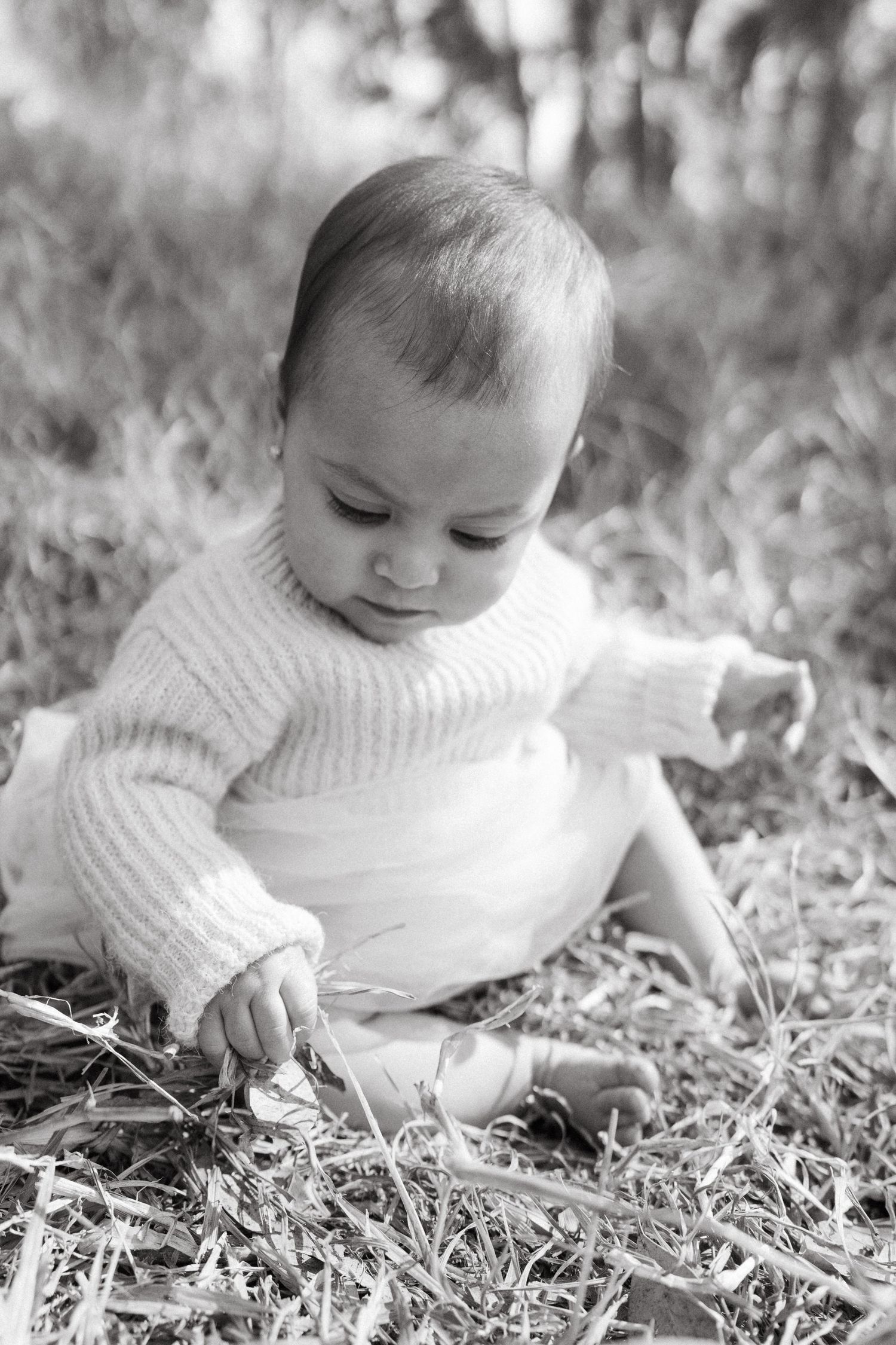 Black and white series of baby in white sweater exploring grass and leaves.