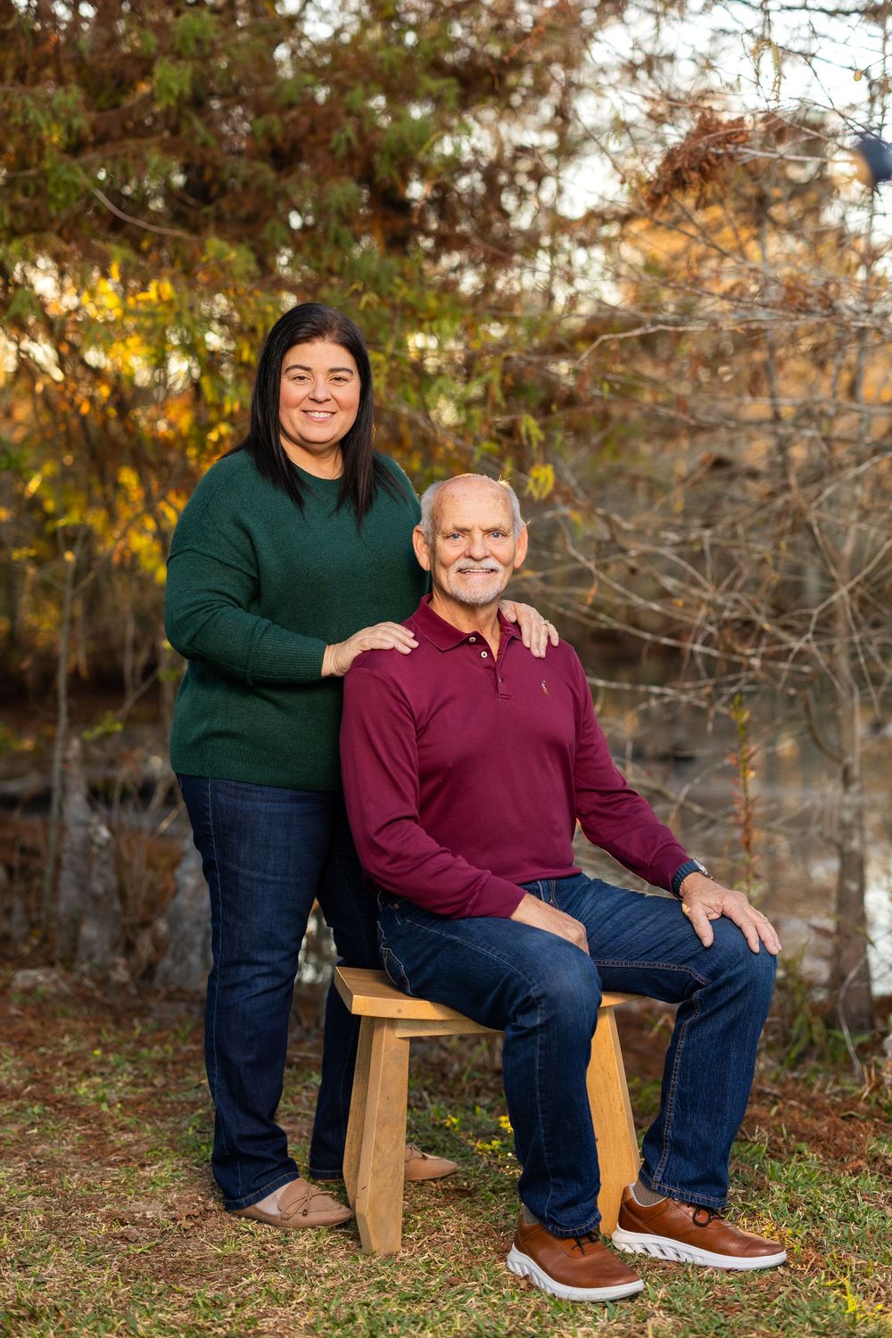 Outdoor portrait of two people on wooden bench with autumn trees in background.
