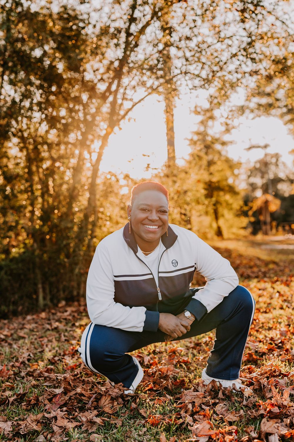 A person in athletic wear crouches and smiles during an autumn photoshoot with fallen leaves.
