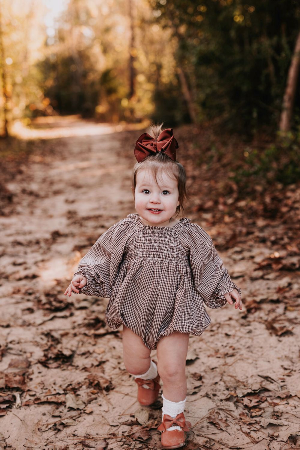 A toddler in a gray dress and bow walks along a leaf-covered forest path.