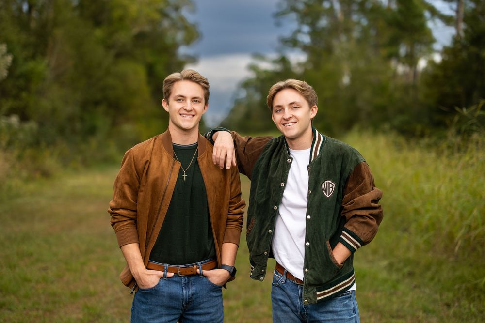 Two people in brown and varsity jackets posing together on a grassy hillside.