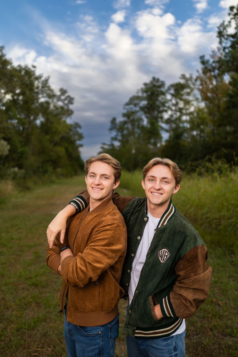 Two people in casual attire posing together outdoors on a grassy hill with trees in the background.