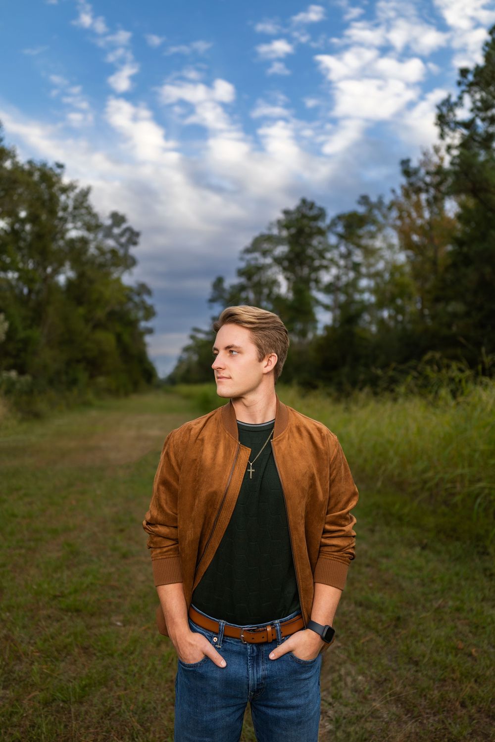 Person in brown jacket and jeans poses casually against scenic outdoor backdrop with cloudy sky.
