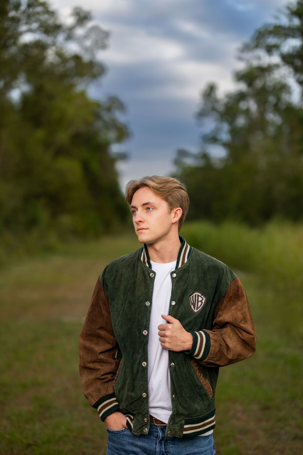 Person wearing varsity jacket poses outdoors on grassy hillside during cloudy day.