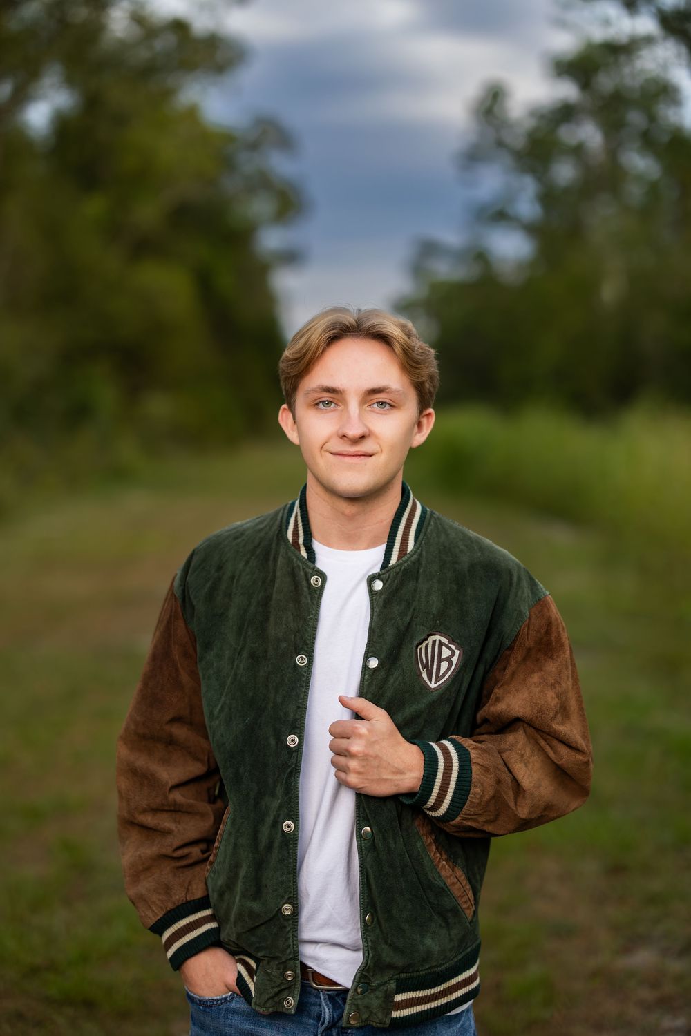Person in varsity jacket and white shirt poses confidently in outdoor nature setting.